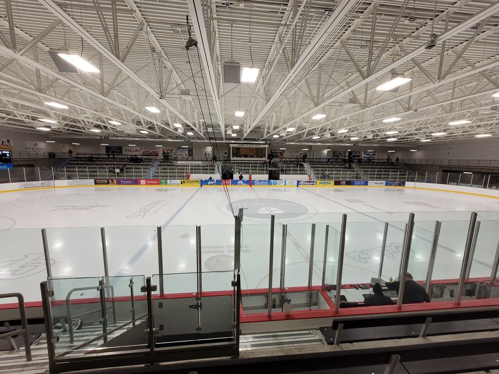 The rink from the stands, a wide sheet seen over the rail with a press-box balcony and a steel-trussed roof above the far wall. — Woodman's Sports and Convention Center
