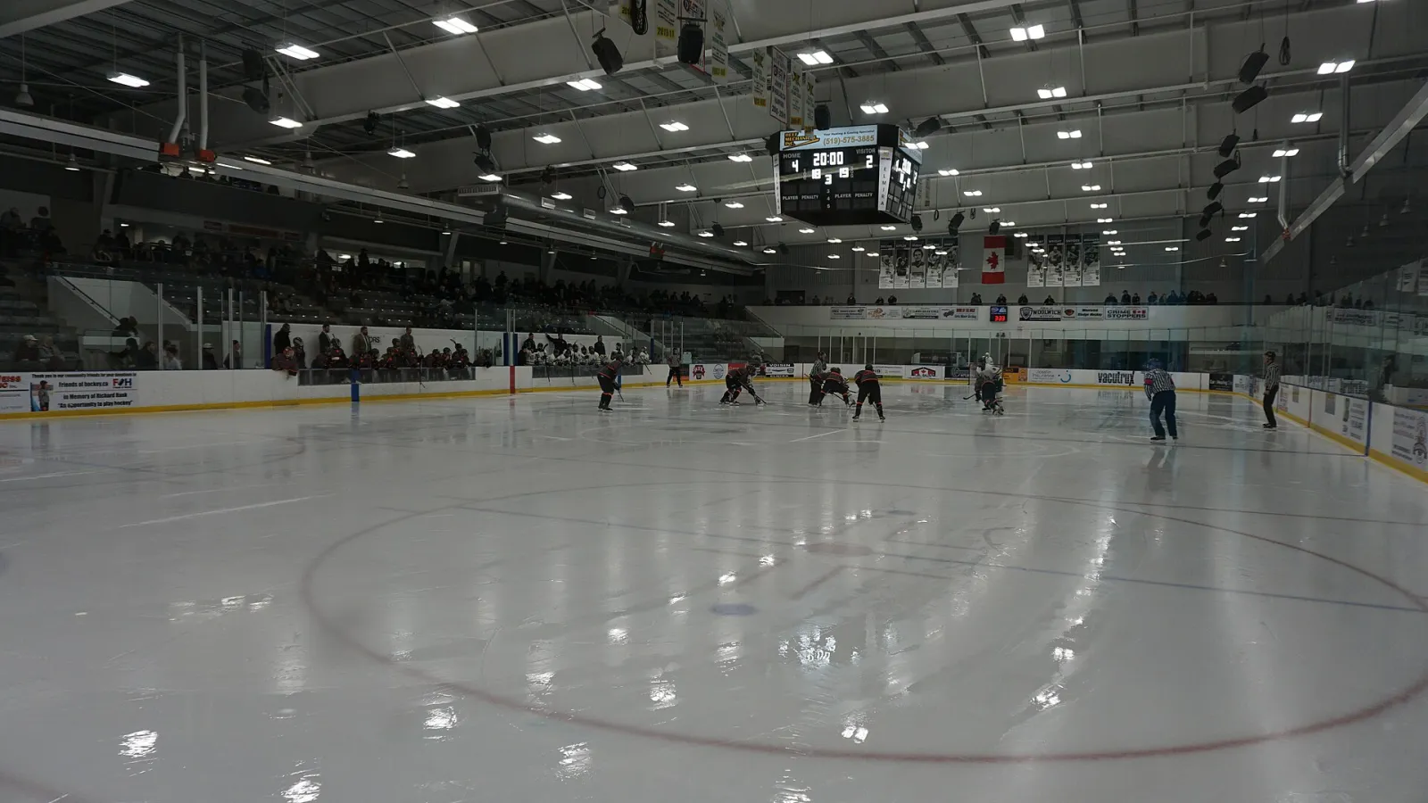 The main rink from the corner, with players on the ice, grey stands and a scoreboard hanging from the white steel roof. — Woolwich Memorial Centre