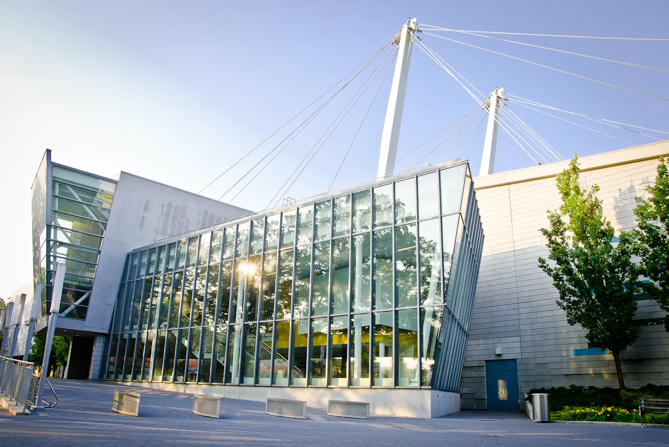 Angled glass curtain-wall entrance pavilion beneath white cable-stayed masts on a sunny evening — World Ice Arena