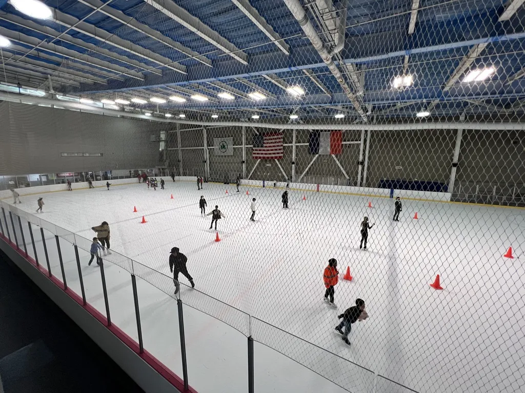 A public session on a wide sheet seen over the rail, skaters spread among orange cones beneath a blue-ceilinged hall with American flags on the far wall. — World Ice Arena