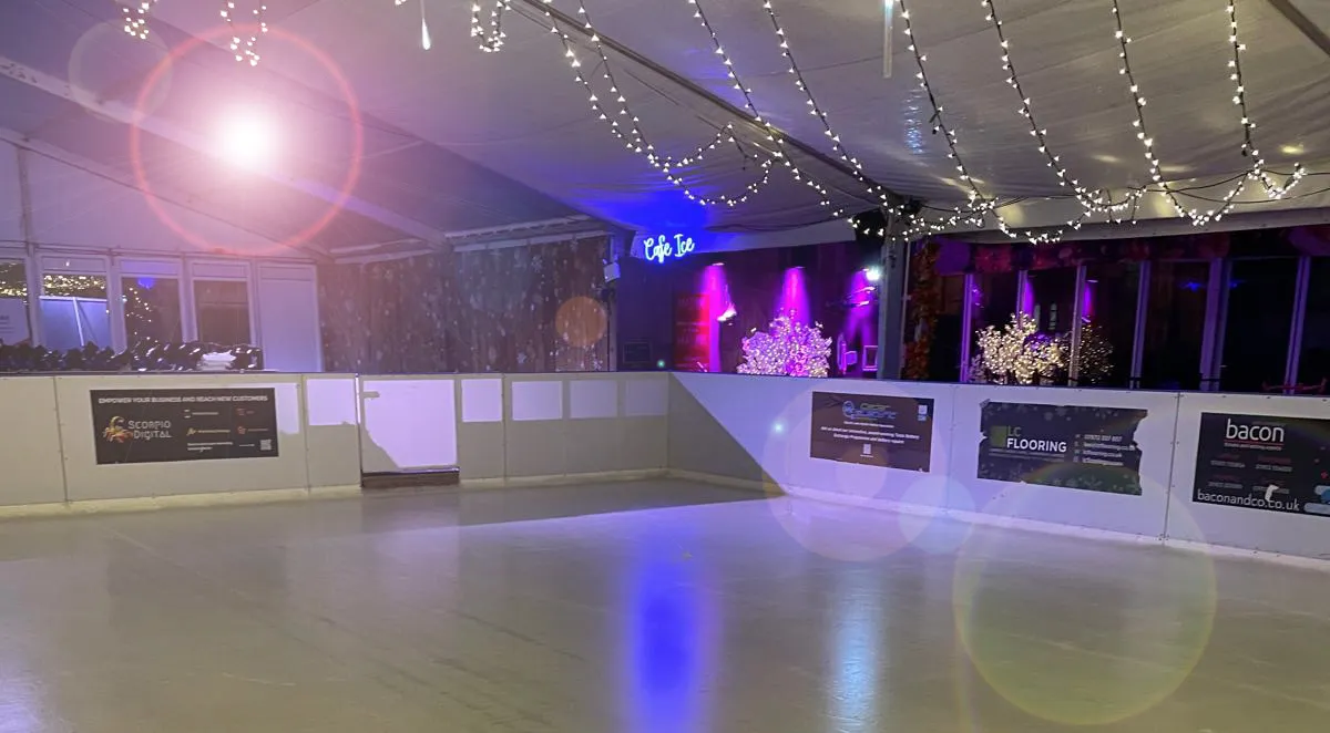 The empty ice sheet inside the Worthing Ice Rink marquee in Steyne Gardens, with string lights, a licensed cafe and sponsor boards along the boards — Worthing Ice Rink