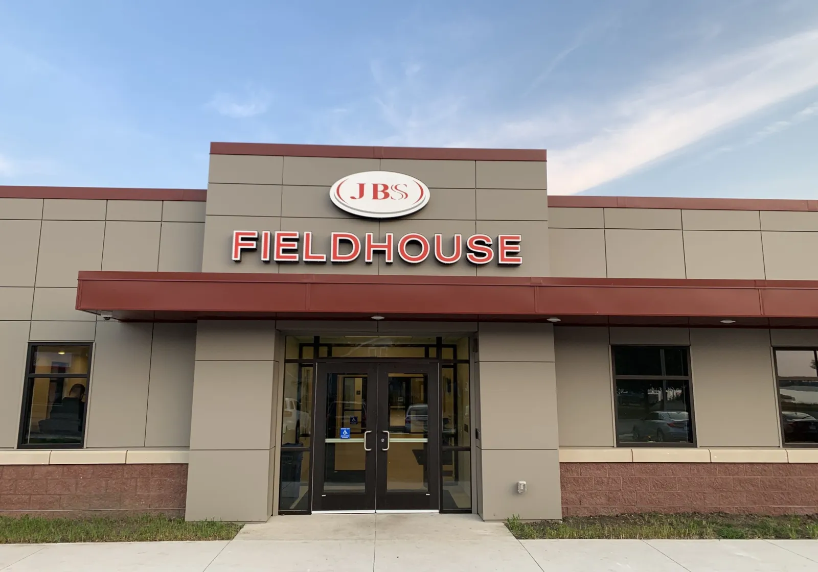 The JBS Fieldhouse entrance at 1600 Stower Drive, the building address the Worthington Arena ice sheet is listed under, with red lettering over a tan panel front and glass doors under a pale blue sky. — Worthington Arena