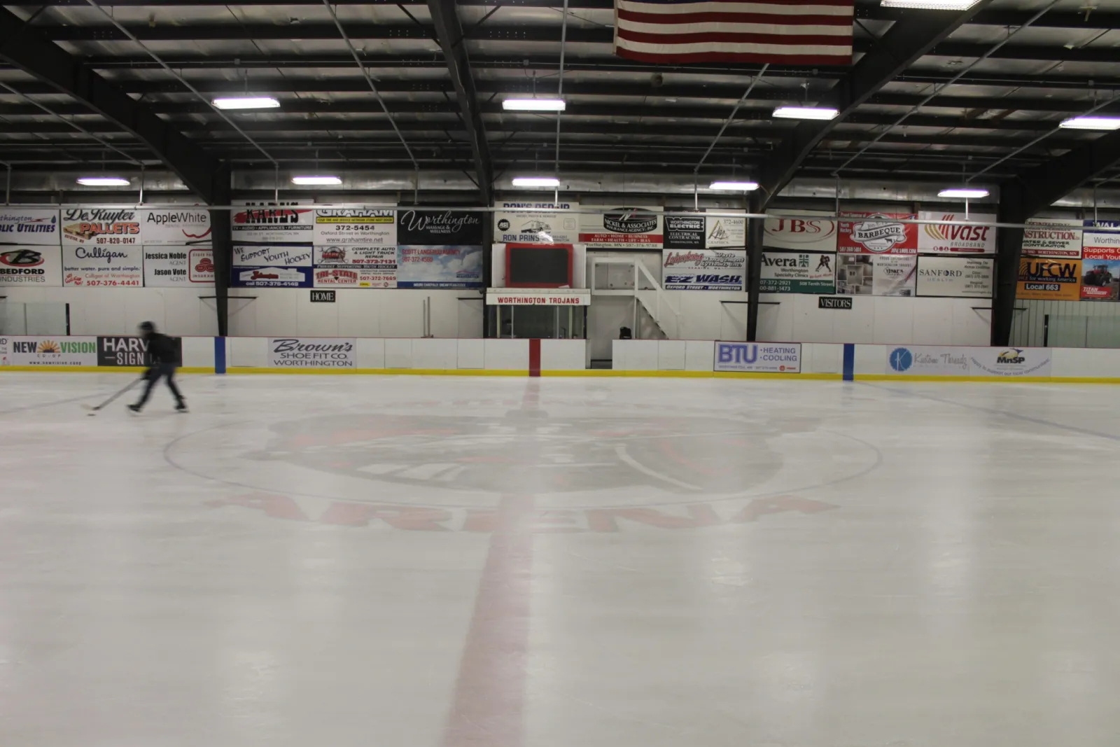 The sheet at centre ice with a single skater at the far boards, sponsor signs on the walls and an American flag under the steel roof. — Worthington Arena