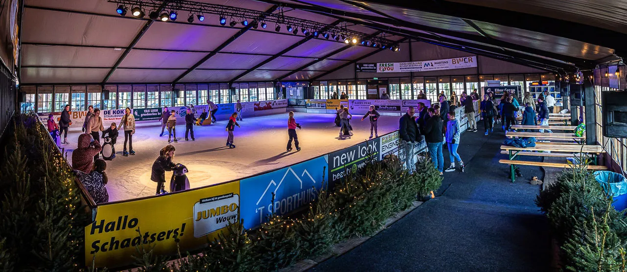 Schaatsers op de overdekte ijsbaan in het wintersportchalet-tentpaviljoen, met de boarding en toeschouwers langs de kant. – Wouw On Ice