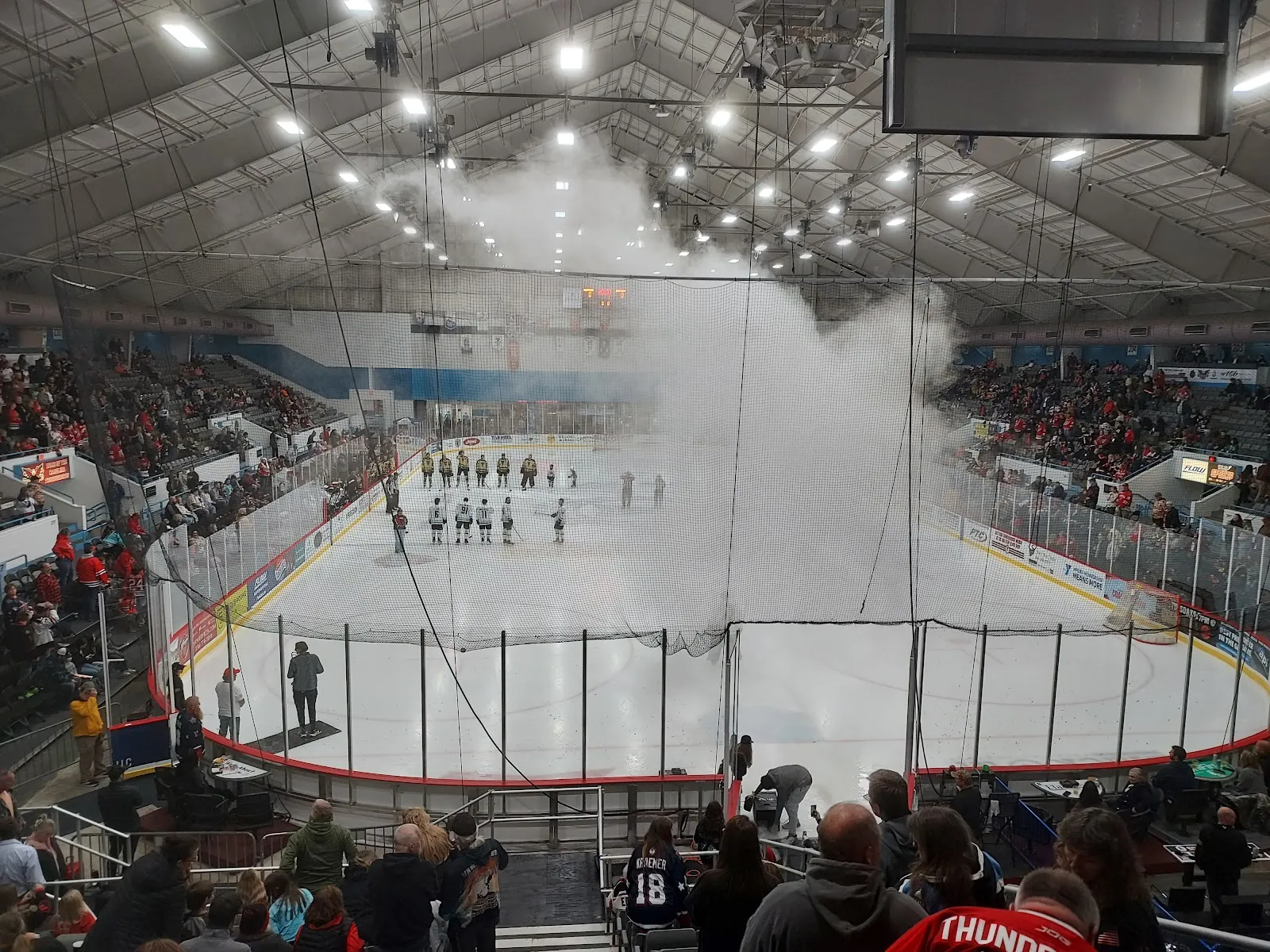 The arena bowl with the ice lit under the roof trusses, teams lined up on the sheet and spectators filling the stands around it. — Winston-Salem Fairgrounds Arena