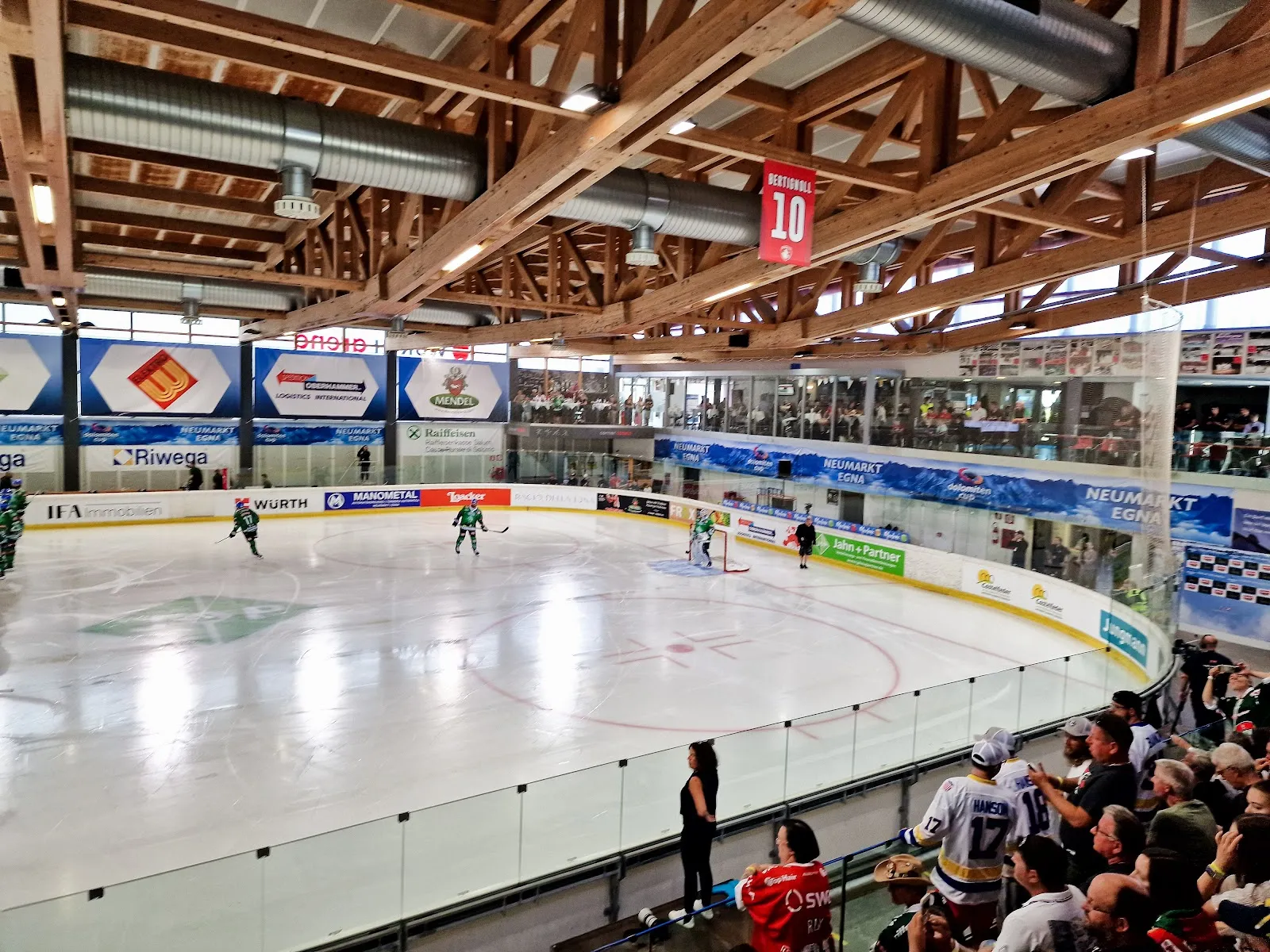 Una partita di hockey su ghiaccio sotto le travi di legno del tetto dell'arena, vista dalla tribuna, con i cartelloni degli sponsor lungo la balaustra e gli spettatori in tribuna e nella galleria superiore. – WürthArena Egna