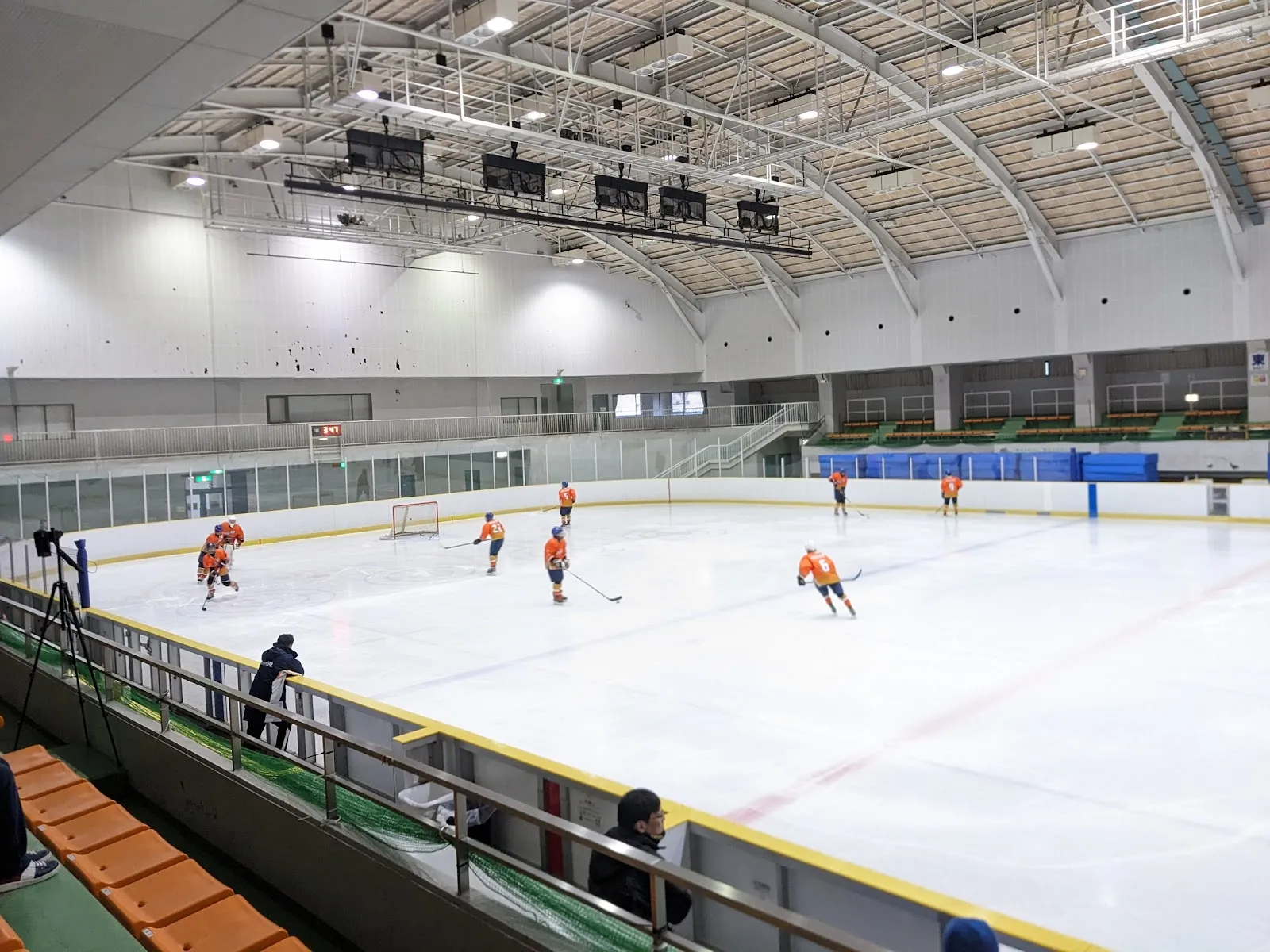 A wide view of the Yamabiko Ice Arena's ice sheet from the stands, with a small group of hockey players spread across the ice under the arched roof — Yamabiko Ice Arena