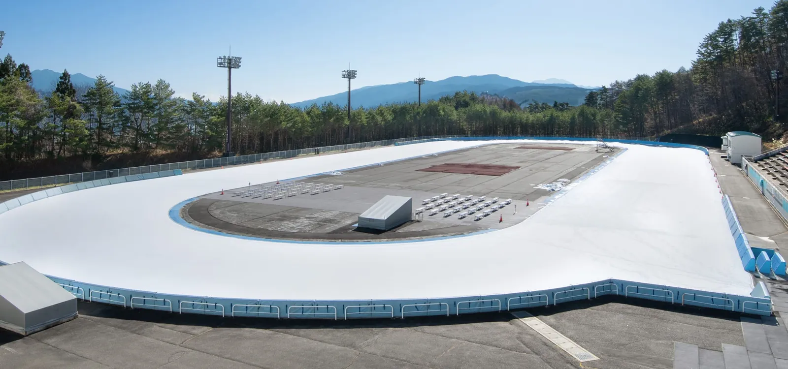 The white 400 m oval seen from the stand on a sunny day, with floodlight masts and wooded hills behind and no skaters on the ice. — Okaya Yamabiko International Skate Center (outdoor oval)