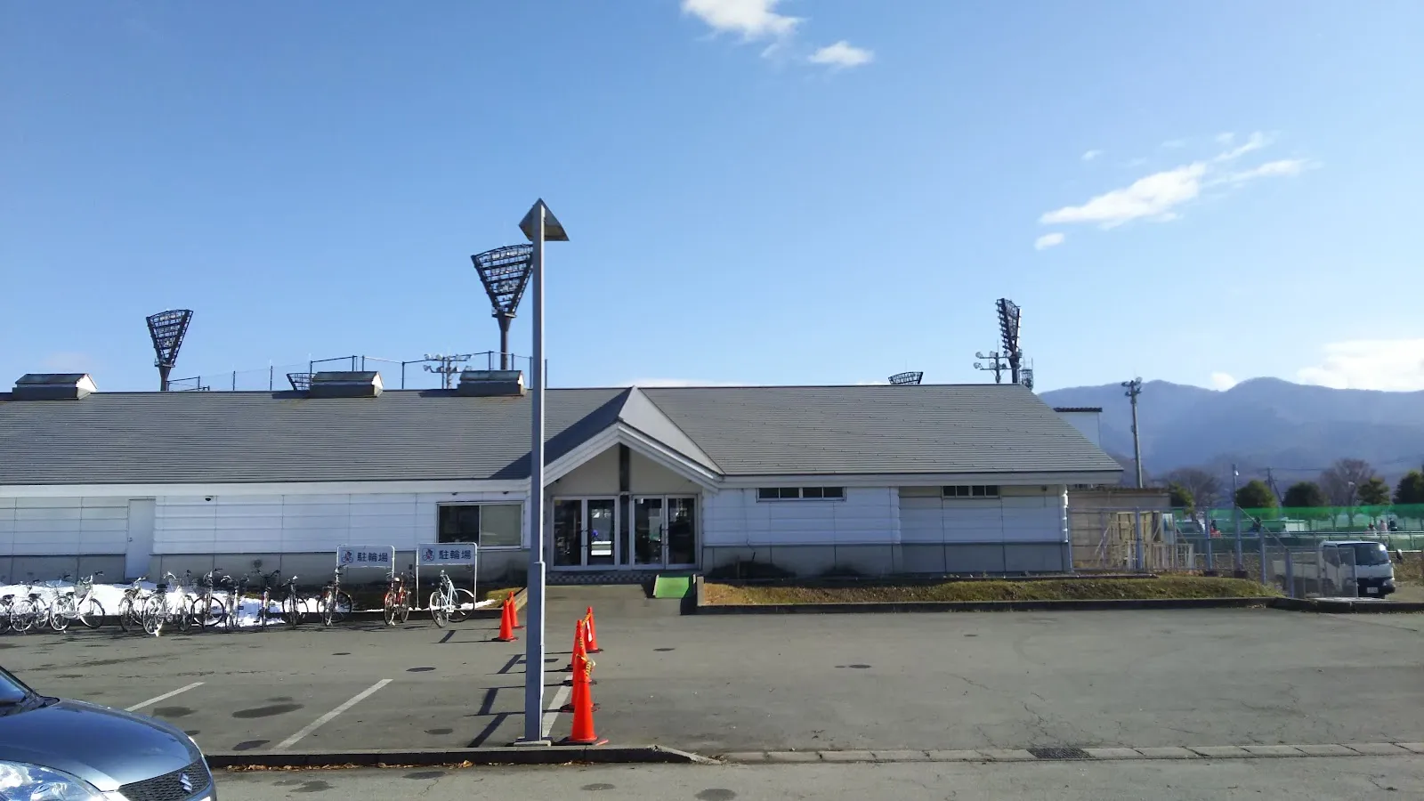 山形市総合スポーツセンターのスケート場側にある白い平屋の入口の建物。屋根の上に照明塔が並び、手前に駐輪場と駐車場がある。 — Yamagata City General Sports Center Skate Rink