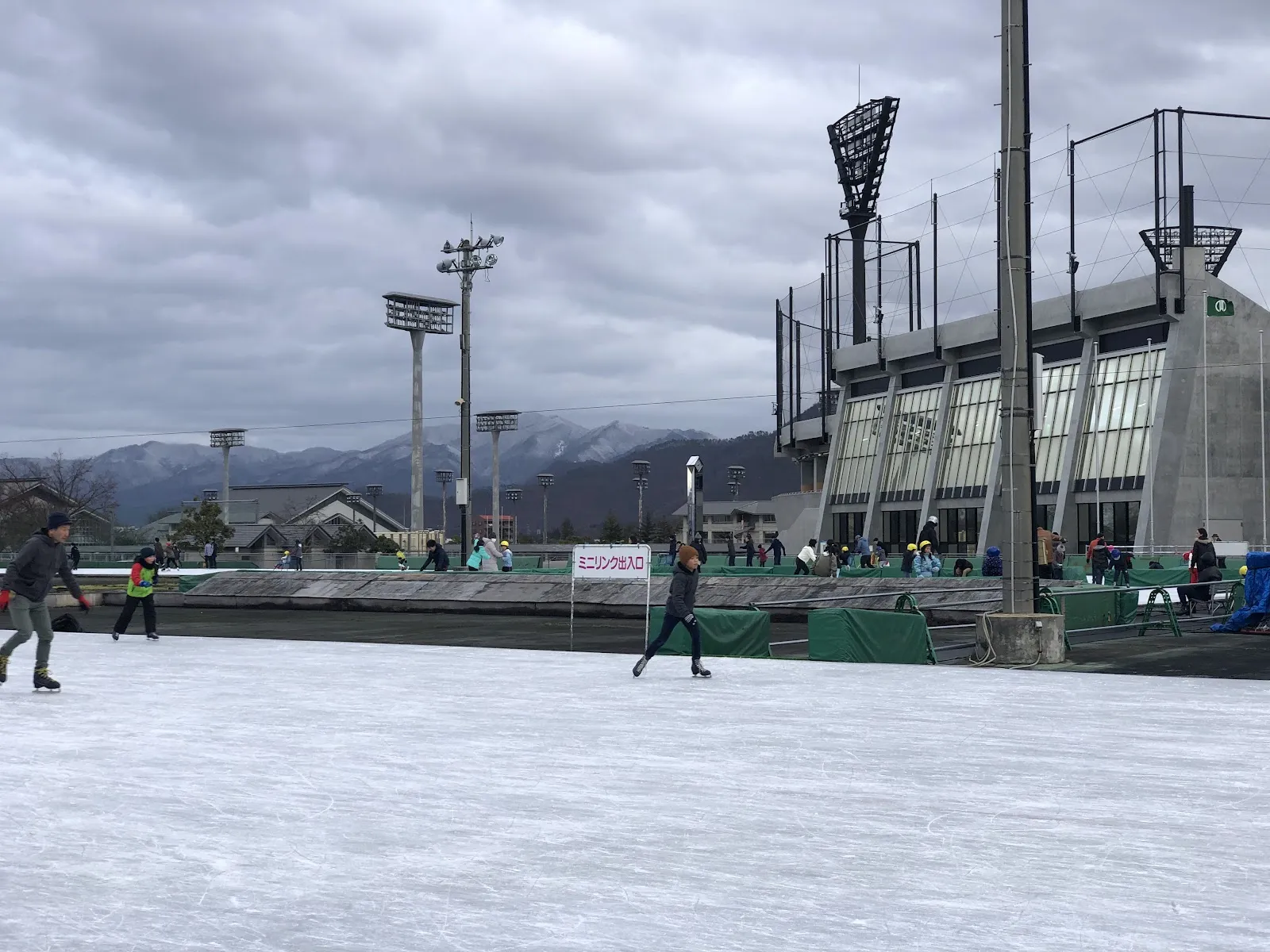 屋外のスピードスケートリンクで滑る人々の写真。ミニリンクの出入口の看板、照明塔、右手にスタンドの建物、背後に雪をかぶった山々が見える。 — Yamagata City General Sports Center Skate Rink