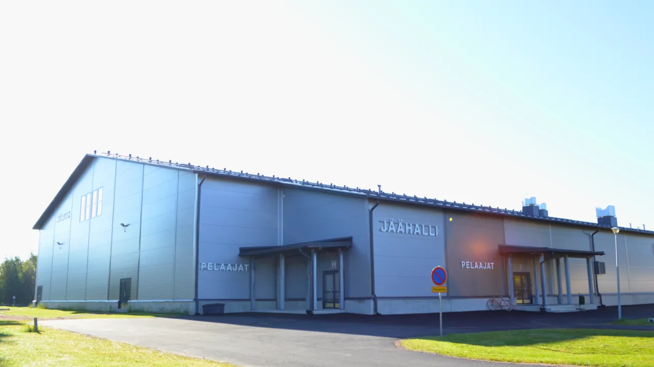 The grey steel-clad Ylivieska ice hall with its JÄÄHALLI sign and two PELAAJAT entrances, seen from the access road across a lawn. – Ylivieskan jäähalli