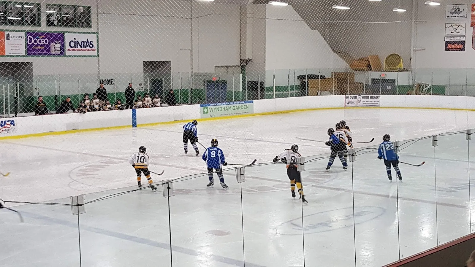 A game seen over the boards, players spread across a wide sheet under a pale steel-framed roof with a grandstand edge at the far end. — York Ice Arena