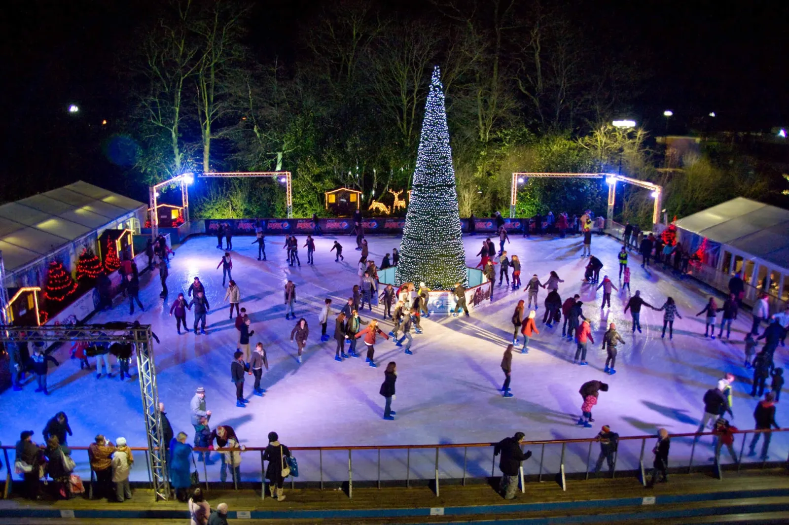 The whole outdoor rink from above at night, skaters circling the ice with a lit Christmas tree at its centre and festive tents around the edge. — Yorkshire's Winter Wonderland Ice Rink