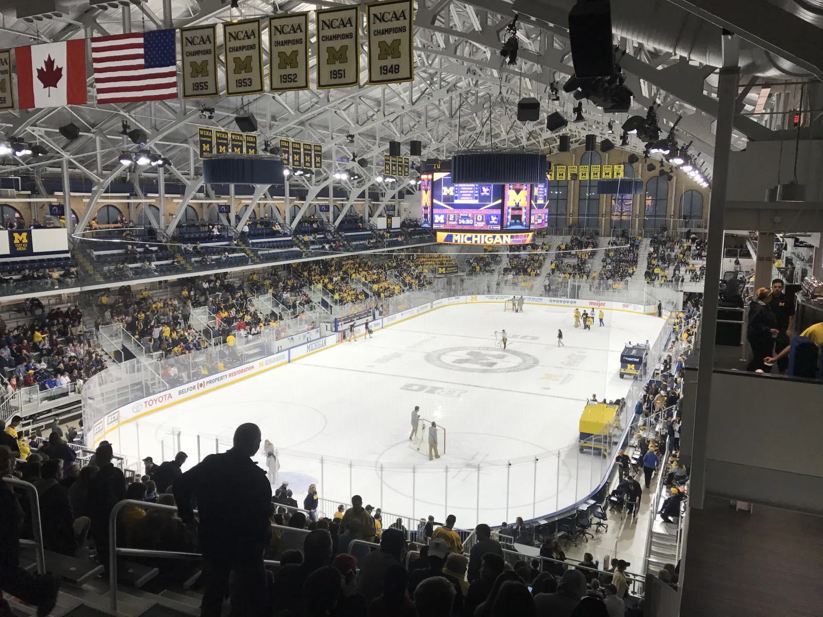 The ice from the upper seating during a Michigan hockey game, with the steel roof trusses, NCAA championship banners, the scoreboard and a crowd in the stands. — Yost Ice Arena