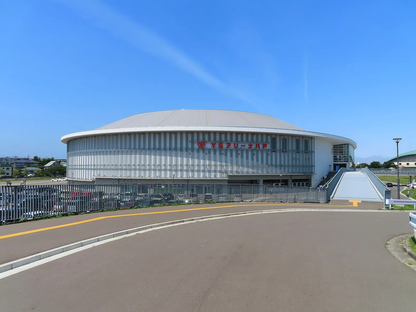 The round white YS Arena Hachinohe seen from the access ramp on a clear day, with the arena's red name sign on the curved wall. — YS Arena Hachinohe (Naganeoku Indoor Skating Rink)