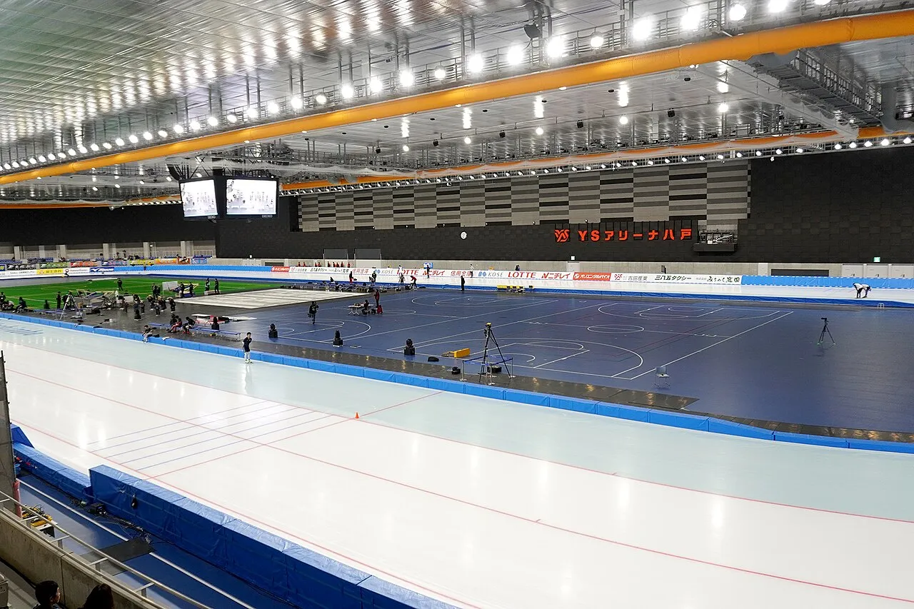 The 400 metre speed-skating oval of YS Arena Hachinohe with a few skaters training, the multi-purpose court inside the oval and the stands and ceiling lighting above. — YS Arena Hachinohe (Naganeoku Indoor Skating Rink)