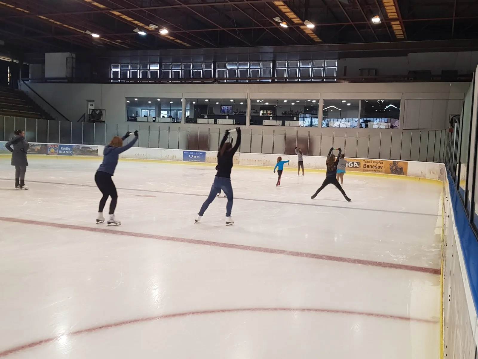 The ice surface of Zimní stadion Benešov from the boards, with a few skaters far out on the ice under the glass-fronted upper gallery — Zimní stadion Benešov
