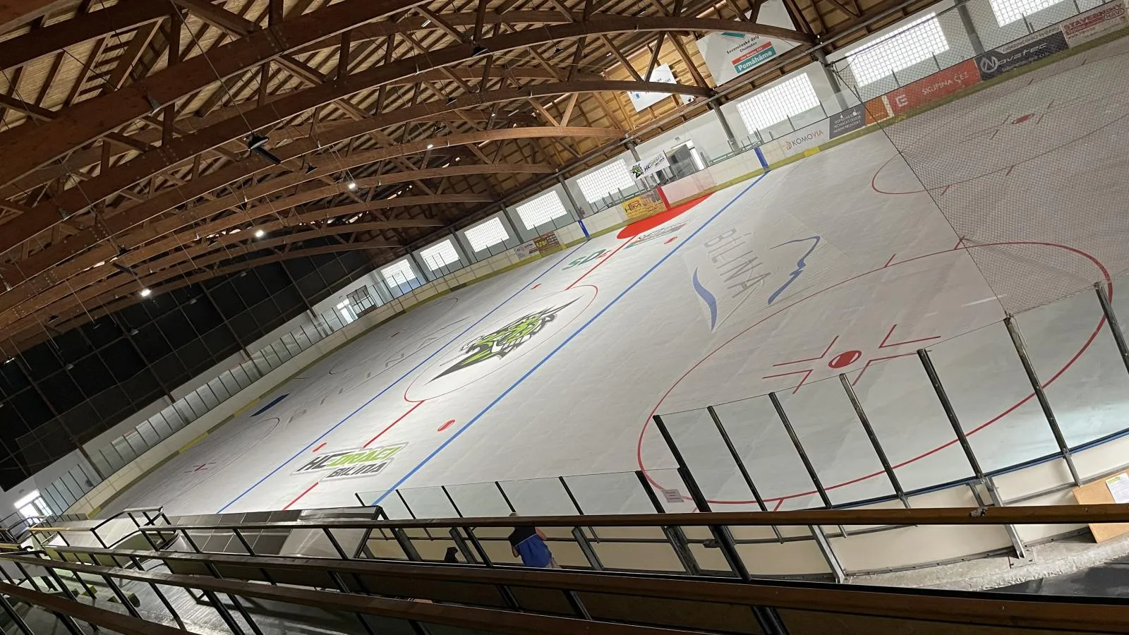 The empty ice sheet of Zimní stadion Bílina with the HC Draci logo at centre ice, seen from the gallery under the timber roof arches — Zimní stadion Bílina