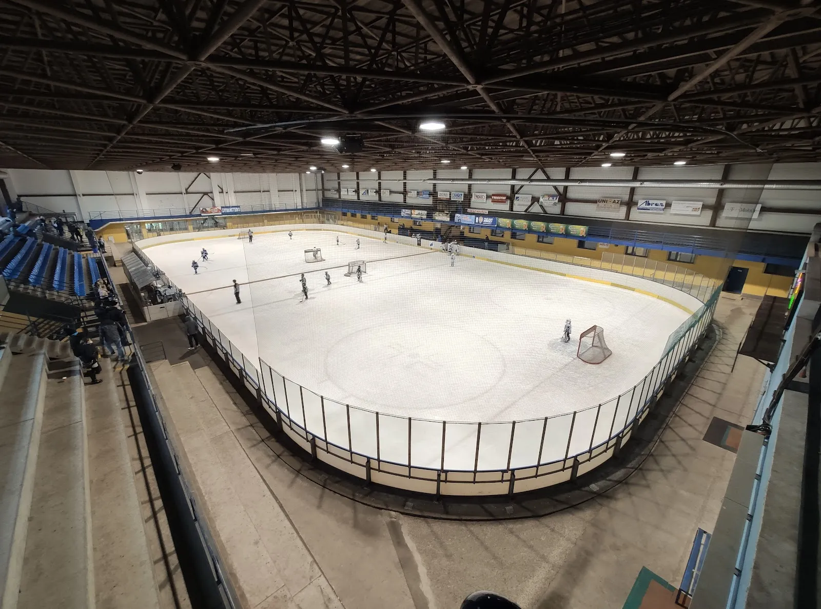 A wide view of the Blansko winter stadium ice from the stand, with the boards, steel roof and a few skaters near the far end. – Zimní stadion Blansko