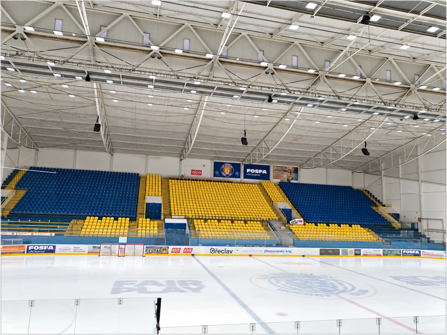 Prázdná ledová plocha Zimního stadionu v Břeclavi pohledem z tribuny, s modrožlutými sedadly a ocelovou střechou nad nimi – Zimní stadion Břeclav