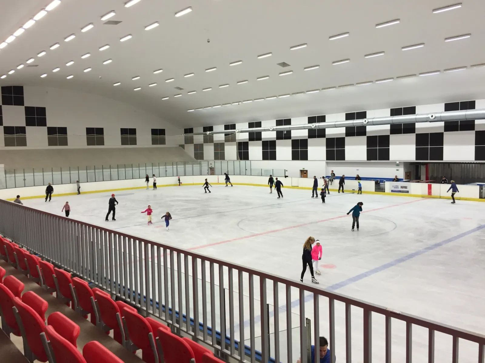 The ice sheet inside the Český Těšín winter stadium with skaters spread across the ice, red seats and the black-and-white chequered wall behind the boards. — Zimní stadion Český Těšín