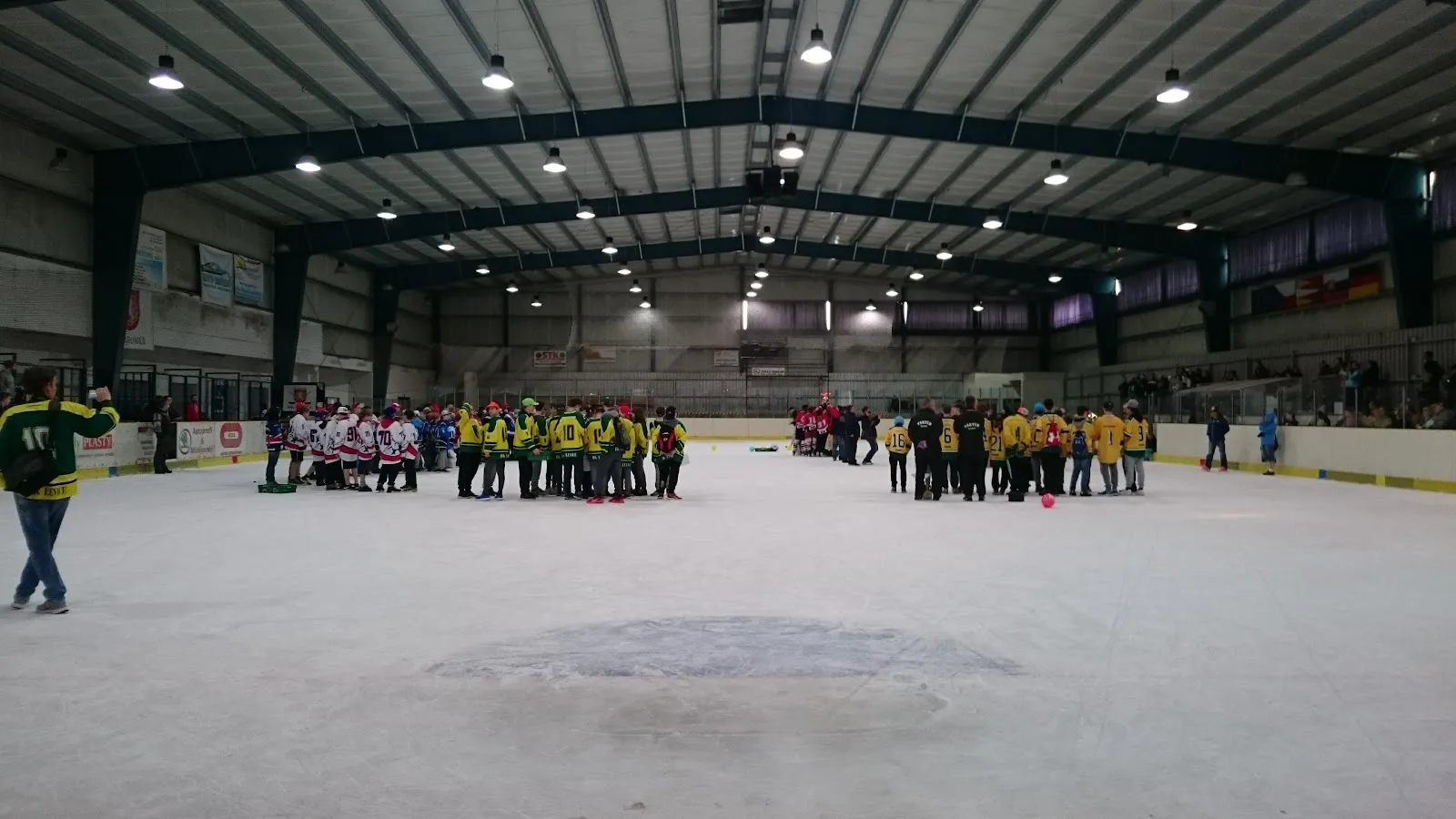 Youth teams in yellow bibs on the Jaroměř winter stadium ice under the dark roof trusses, with the boards behind. — Zimní stadion Jaroměř