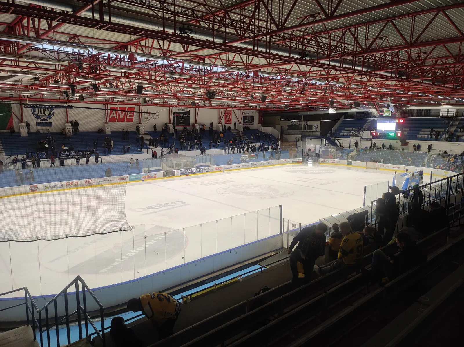 The ice of the Kolín winter stadium during a hockey match, seen from the stands, with spectators on the terraces and the red steel roof trusses above. — Zimní stadion Kolín