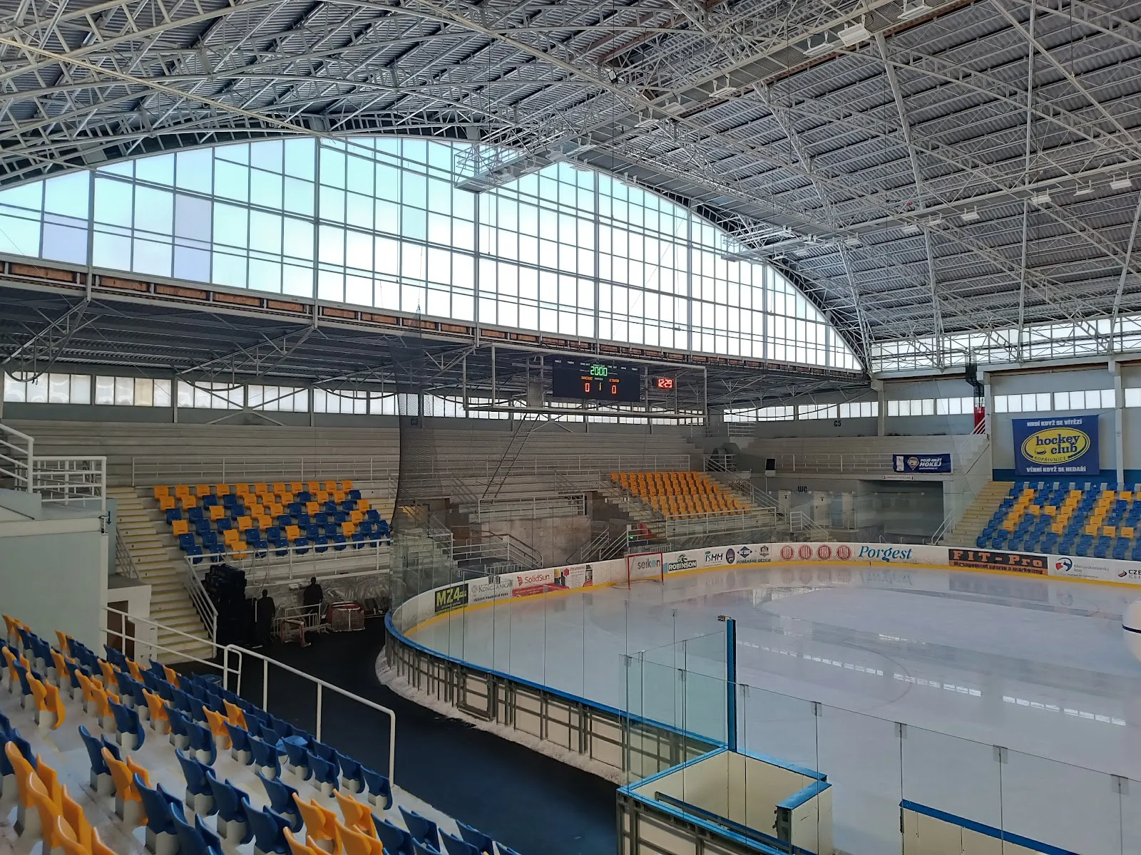 Ledová plocha zimního stadionu v Kopřivnici z tribuny pod obloukovou ocelovou střechou a proskleným štítem, s prázdnými místy k sezení kolem kluziště – Zimní stadion Kopřivnice