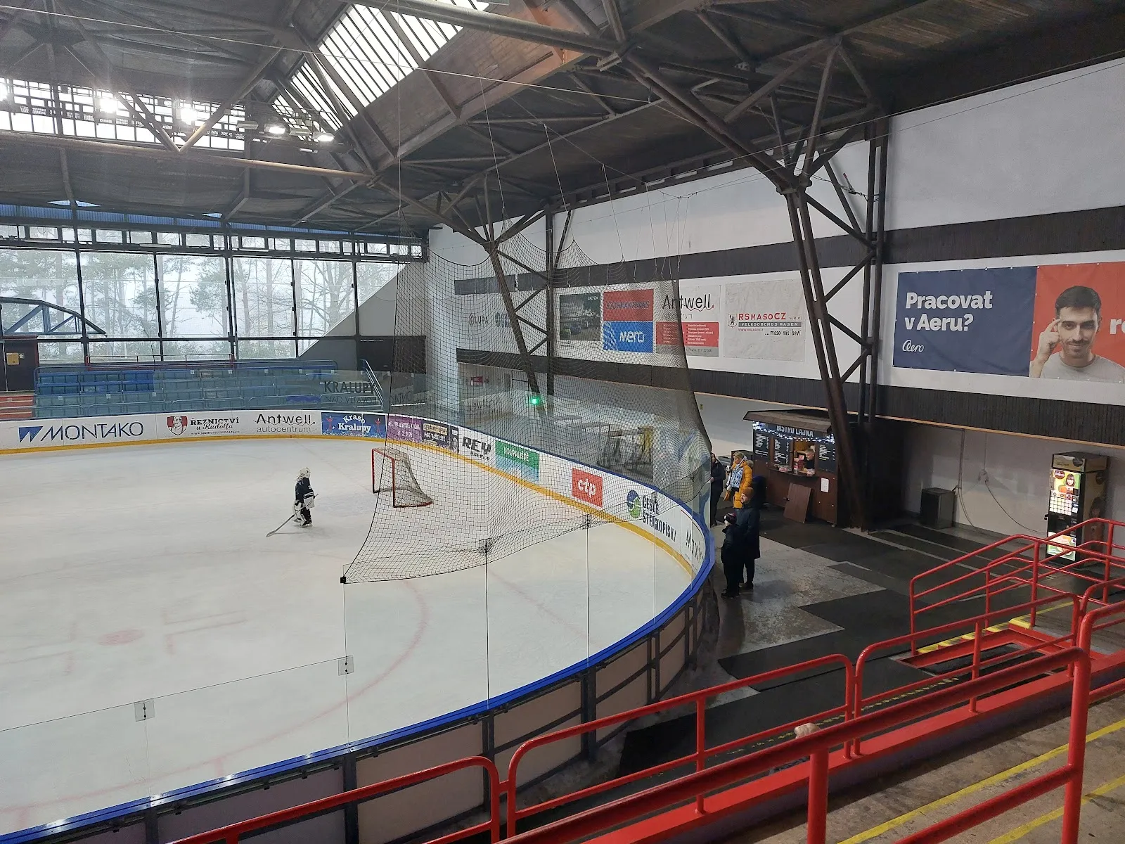 The ice at Kralupy nad Vltavou winter stadium seen from the stands, with the curved blue boards, the steel roof frame, a kiosk and one player small on the ice — Zimní stadion Kralupy nad Vltavou