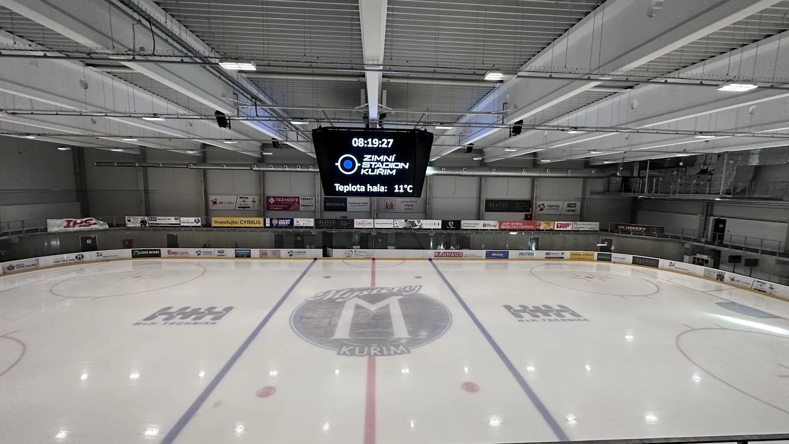 Prázdná ledová plocha pod střechou s ocelovou konstrukcí, s logem klubu uprostřed ledu, videotabulí a mantinely s reklamními panely. – Zimní stadion Kuřim