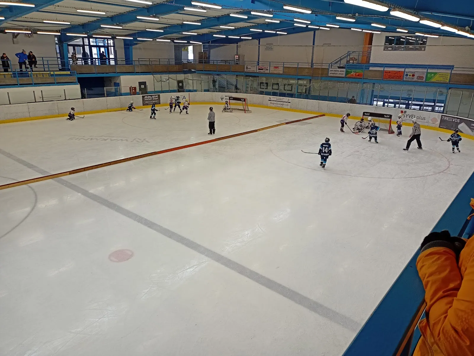 Youth hockey players spread across the Lomnice ice stadium rink, with the boards and a covered stand behind. – Zimní stadion Lomnice nad Popelkou