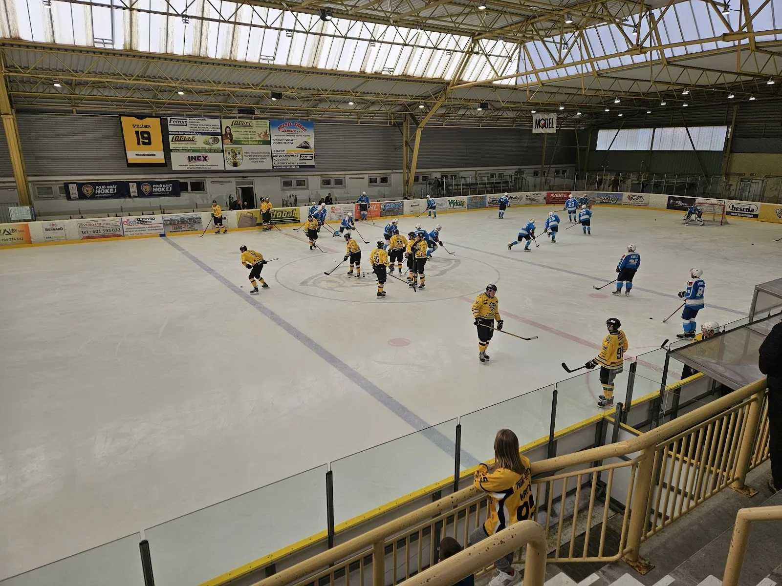A game on the ice of the Moravské Budějovice winter stadium, seen from the stands over the boards, with the hall roof and the far-side stand behind the players. — Zimní stadion Moravské Budějovice