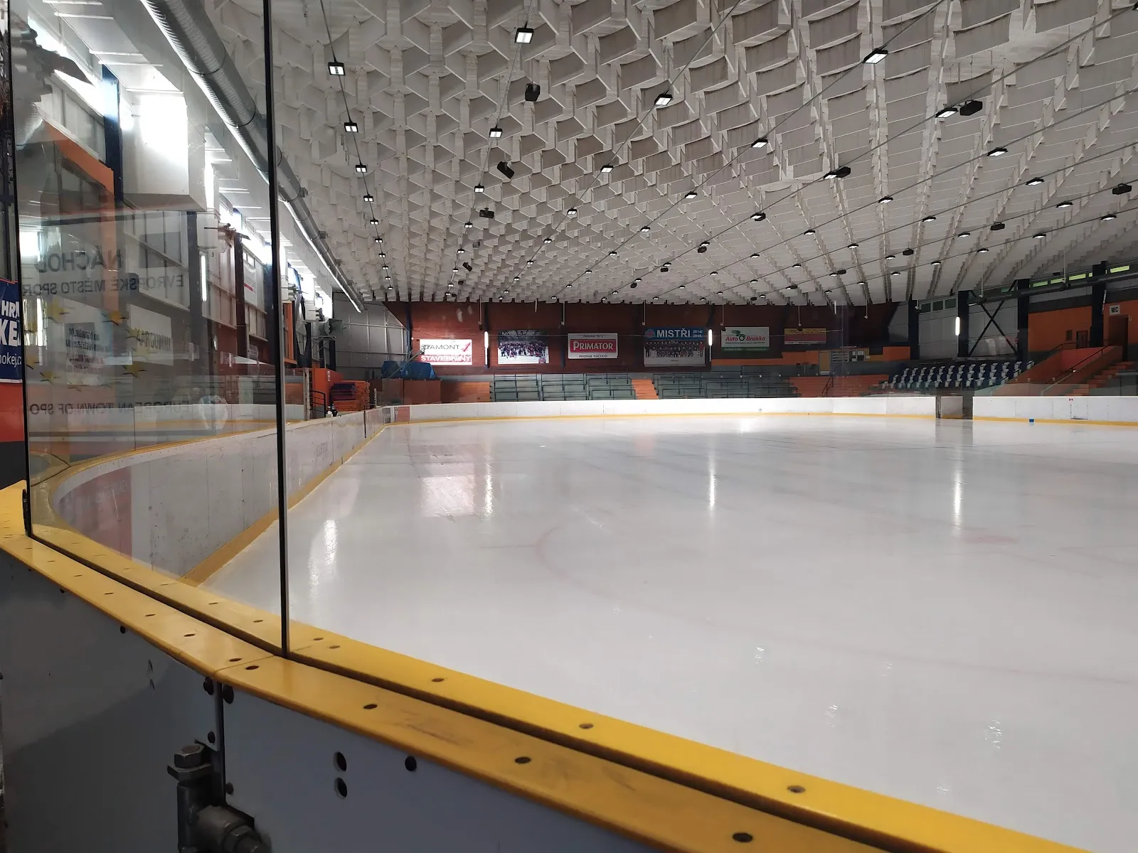 Empty ice at the Náchod winter stadium seen from the boards, with the yellow board rim, a glass panel and the ribbed ceiling of the hall, with the stands behind — Zimní stadion Náchod
