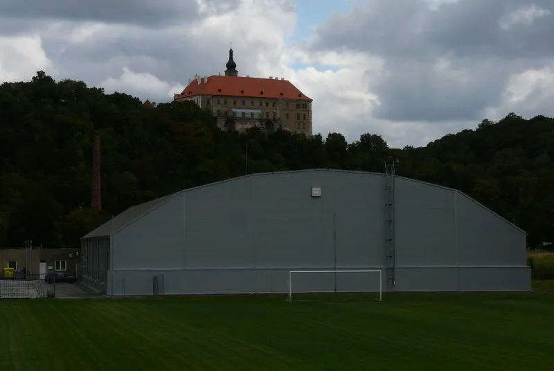 The grey arched hall of the Náměšť nad Oslavou winter stadium seen across a football pitch, with the red-roofed castle on the wooded hill rising behind it. — Zimní stadion Náměšť nad Oslavou