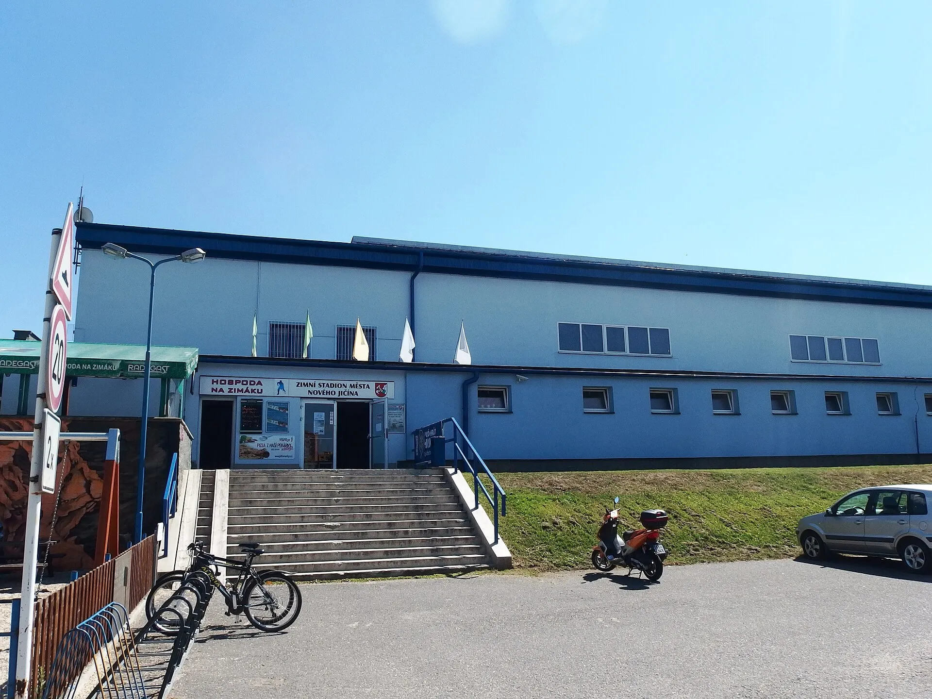 Modré průčelí Zimního stadionu města Nového Jičína se vstupním schodištěm, nápisem Hospoda na Zimáku a vlajkami nad vchodem – Zimní stadion Nový Jičín