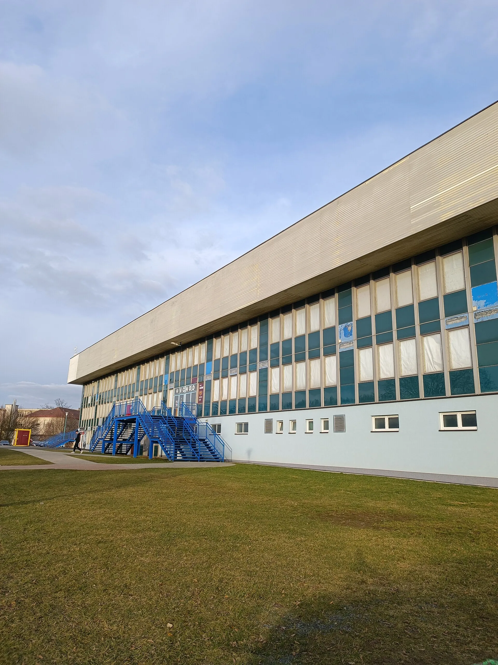 The long side of the Zimní stadion in Pelhřimov with its flat roof, the glazed upper floor and the blue outside staircase — Zimní stadion Pelhřimov