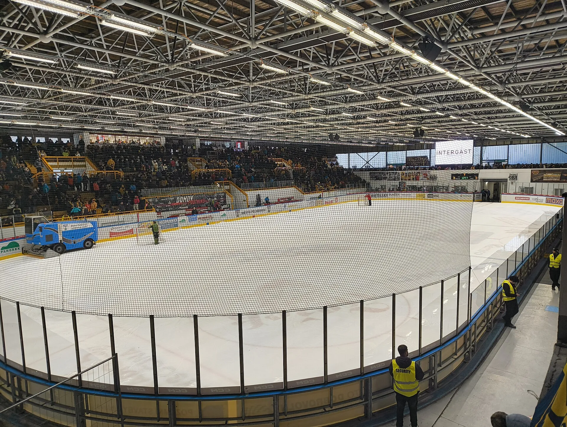 A hockey match at the Zimní stadion in Pelhřimov, seen from the stand with the crowd, the ice resurfacer on the sheet and the steel roof above — Zimní stadion Pelhřimov