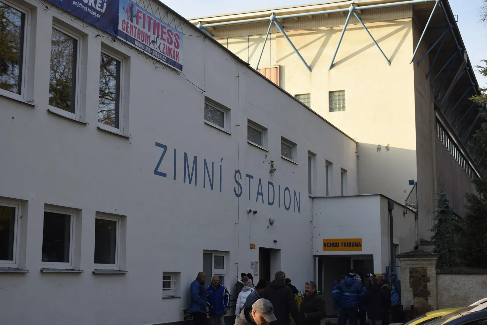 Uliční průčelí zimního stadionu v Písku s nápisem „Zimní stadion“ a vchodem na tribunu, před nímž se shromažďují fanoušci. – Zimní stadion Písek