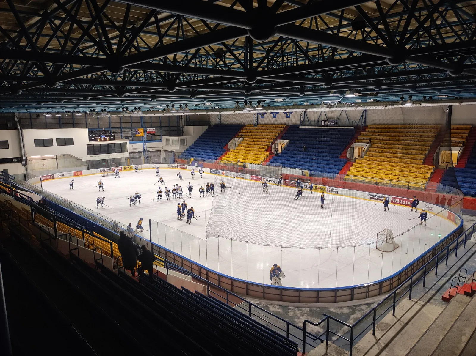 Hokejový trénink na ledě zimního stadionu v Písku při pohledu z tribuny, s ocelovými příhradovými nosníky střechy nahoře a modrožlutými sedadly vzadu. – Zimní stadion Písek