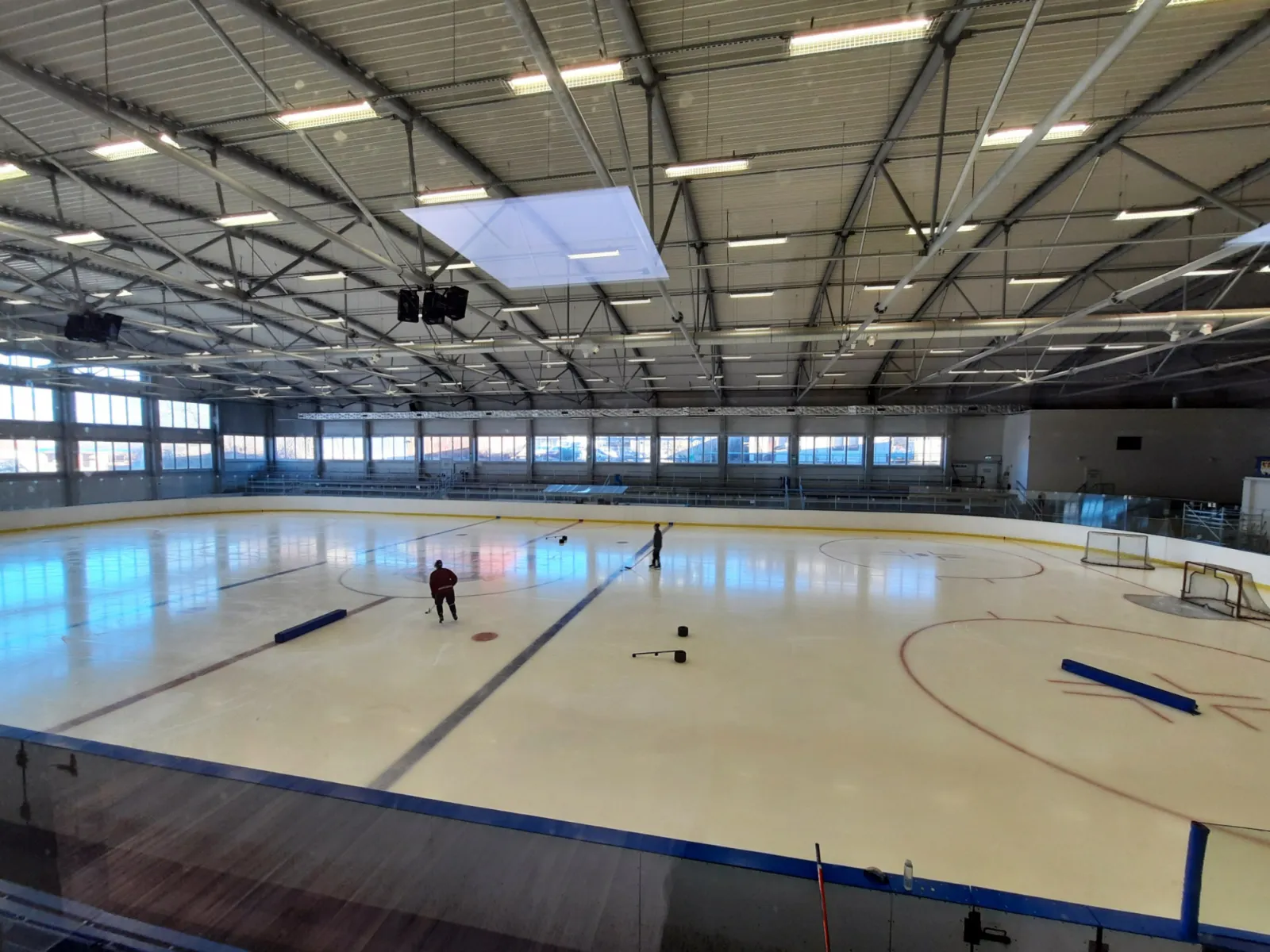 The ice surface of Zimní stadion Poděbrady seen from the stands, with the steel roof trusses and the long side windows; a few people are far away on the ice — Zimní stadion Poděbrady