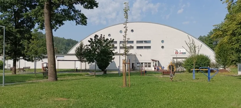 Bílá klenutá střecha zimního stadionu v Rožnově pod Radhoštěm při pohledu přes trávník mezi stromy v letním dni. – Zimní stadion Rožnov pod Radhoštěm