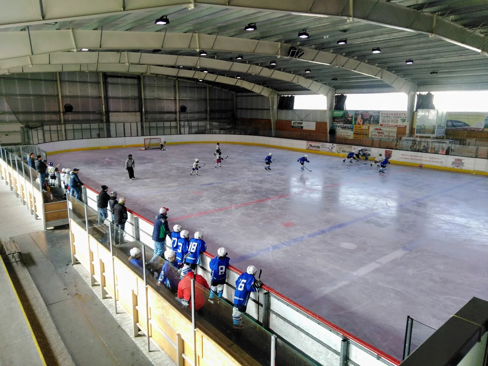 Youth hockey on the ice sheet of the winter stadium in Rychnov nad Kněžnou, seen from the stand with the team bench at the boards — Zimní stadion Rychnov nad Kněžnou