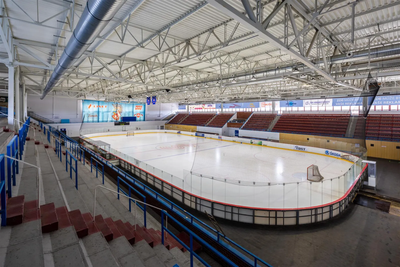 The empty ice sheet of the Zimní stadion in Slaný seen from the stand, with the red stand sections and the steel roof above — Zimní stadion Slaný