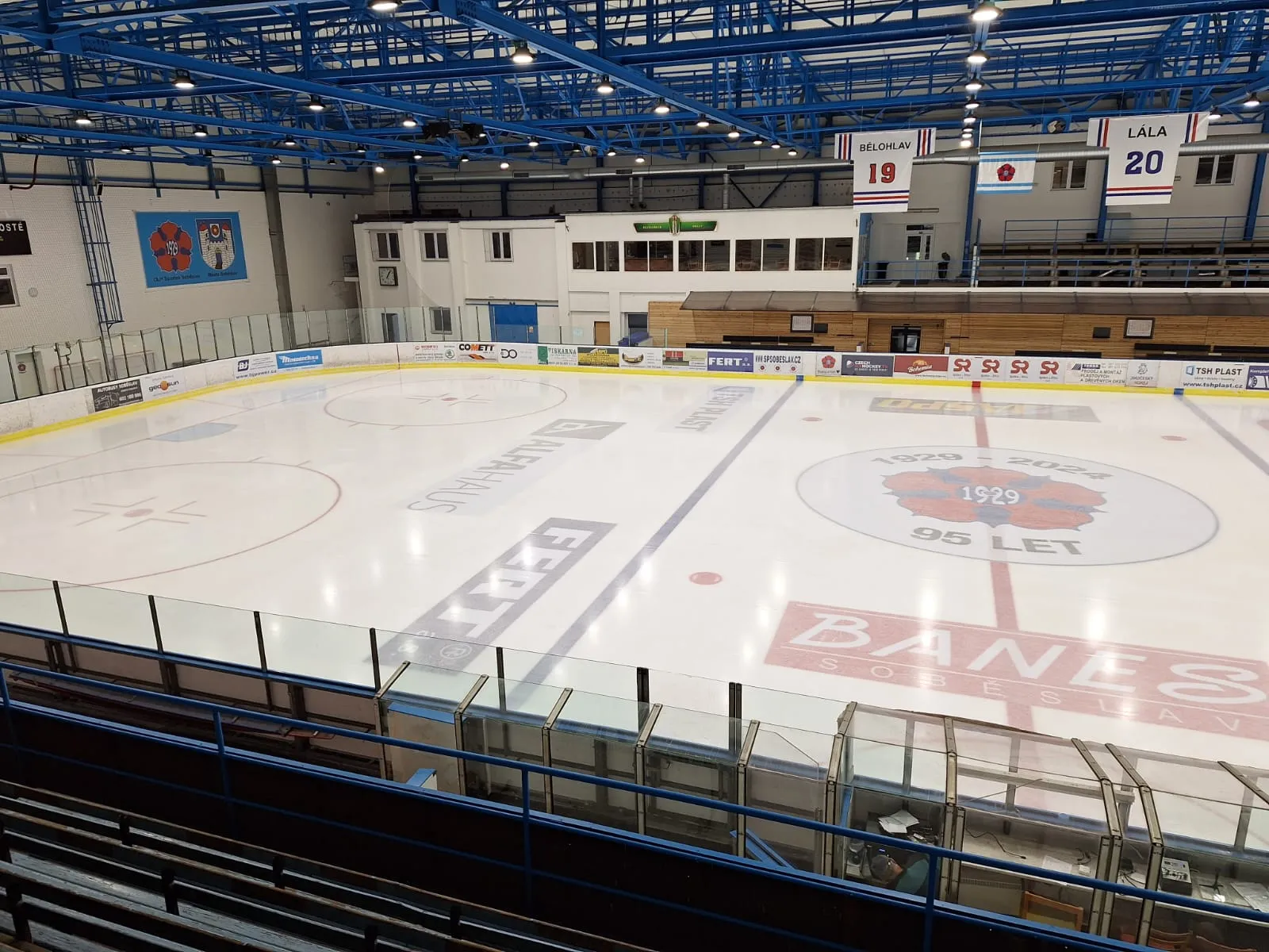 The empty ice sheet of the Soběslav winter stadium seen from the stands, with the 95-year club crest at centre ice, blue roof trusses overhead and the boards lined with advertising. — Zimní stadion Soběslav
