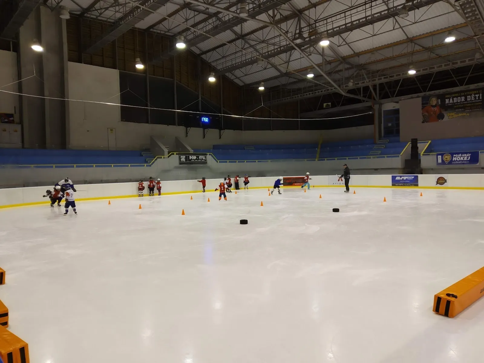 Players and skaters on the ice of the Studénka winter stadium, seen from the boards across the sheet under the roof lighting. — Zimní stadion Studénka