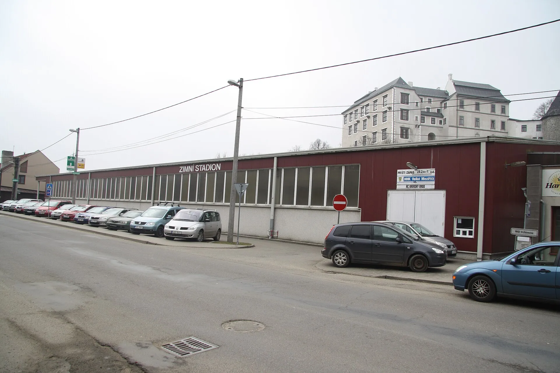Dlouhé uliční průčelí zimního stadionu ve Velkém Meziříčí s názvem stadionu na stěně a auty zaparkovanými podél silnice – Zimní stadion Velké Meziříčí