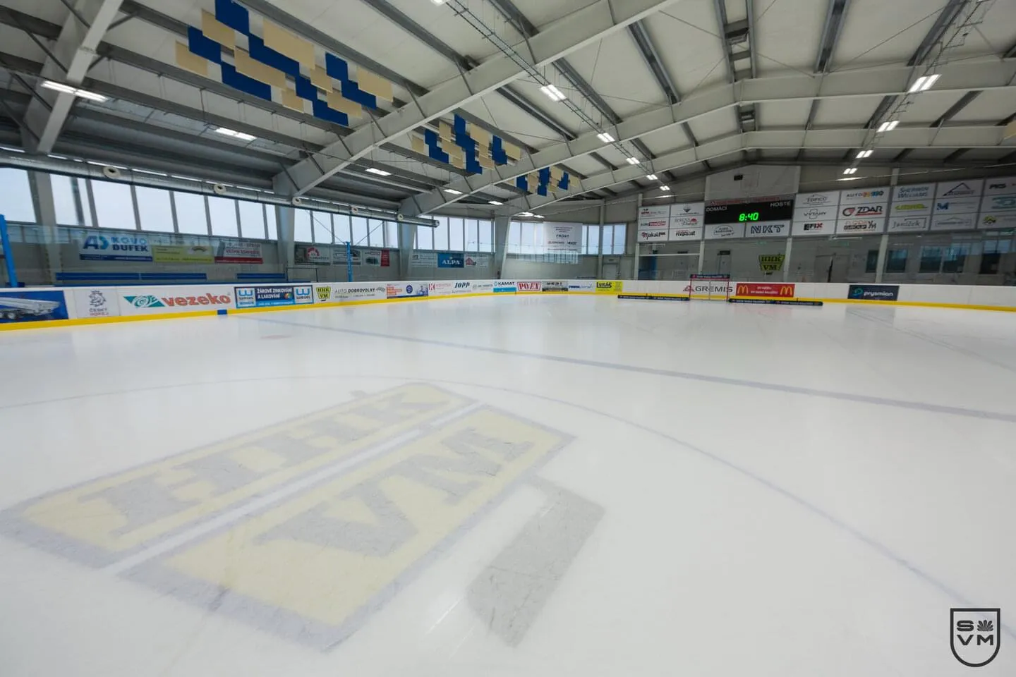 Prázdná ledová plocha zimního stadionu ve Velkém Meziříčí pod ocelovou střechou, s logem HHK VM na ledě – Zimní stadion Velké Meziříčí