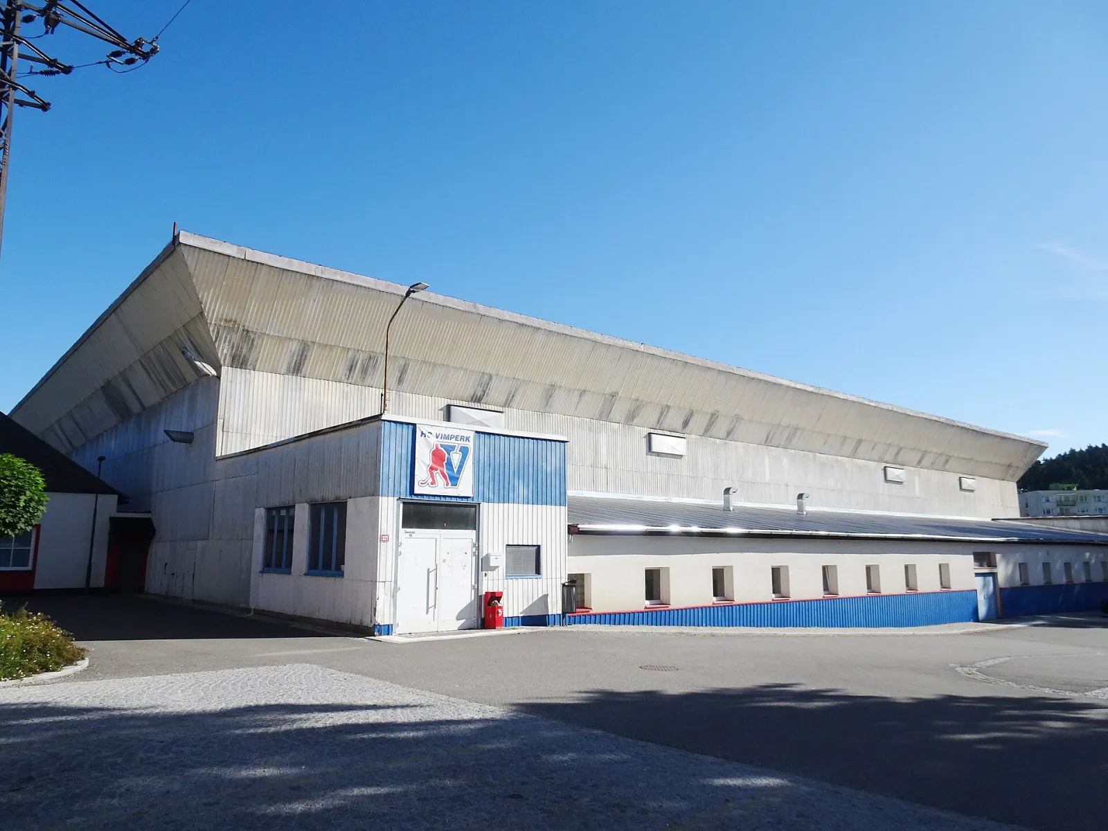 The Vimperk winter stadium seen from the car park: a long grey steel-clad hall with a sloping roof, a blue-and-white entrance block carrying the club sign, and a blue-trimmed lower wing. – Zimní stadion Vimperk