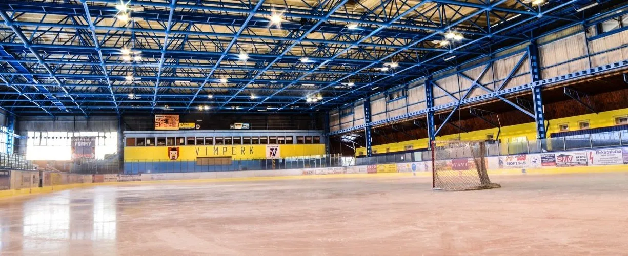 The empty ice sheet of the Vimperk winter stadium under a blue steel roof, with a goal on the right, the yellow-and-blue stand wall marked VIMPERK at the far end and advertising boards along the sides. – Zimní stadion Vimperk