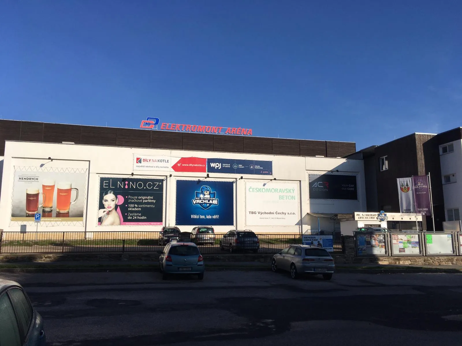 The street front of the Vrchlabí winter stadium under its red 'Elektromont aréna' sign, with advertising banners on the white wall and the club's ticket hut at the right. — Zimní stadion Vrchlabí (Elektromont aréna)